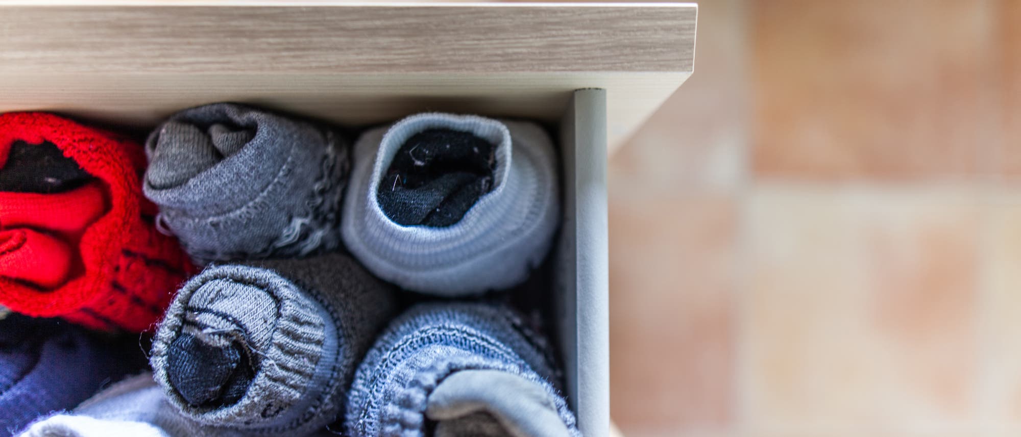 Eine Schublade mit mehreren Paaren gerollter Socken in verschiedenen Farben, darunter Rot, Grau und Blau. Die Socken sind ordentlich nebeneinander angeordnet. Die Schublade ist aus hellem Holz, und der Hintergrund zeigt einen unscharfen Bodenbelag in warmen T&ouml;nen.