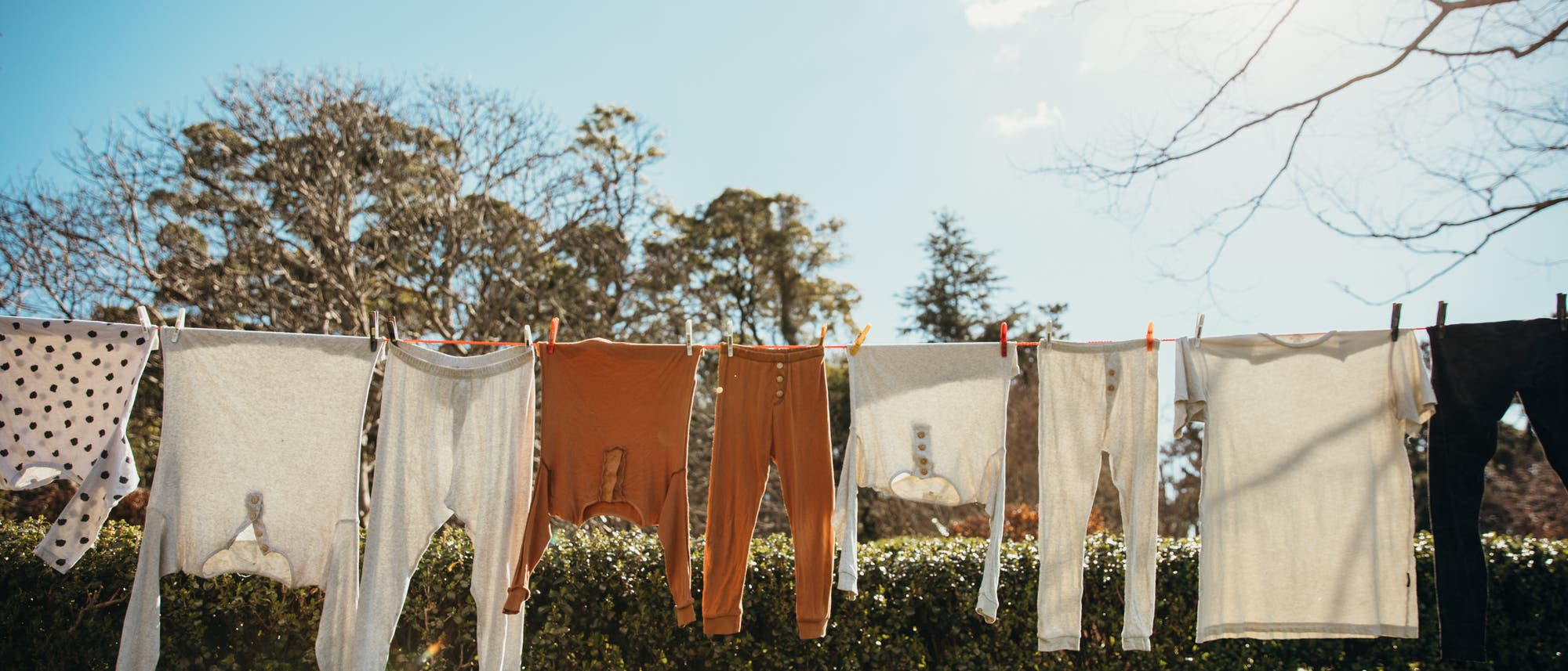 Eine Wäscheleine im Freien, an der verschiedene Kleidungsstücke hängen, darunter weiße und orangefarbene Oberteile und Hosen. Im Hintergrund sind Bäume und ein klarer blauer Himmel zu sehen. Die Szene vermittelt einen sonnigen Tag. Eine Wäscheleine im Freien, an der verschiedene Kleidungsstücke hängen, darunter weiße und orangefarbene Oberteile und Hosen. Im Hintergrund sind Bäume und ein klarer blauer Himmel zu sehen. Die Szene vermittelt einen sonnigen Tag.