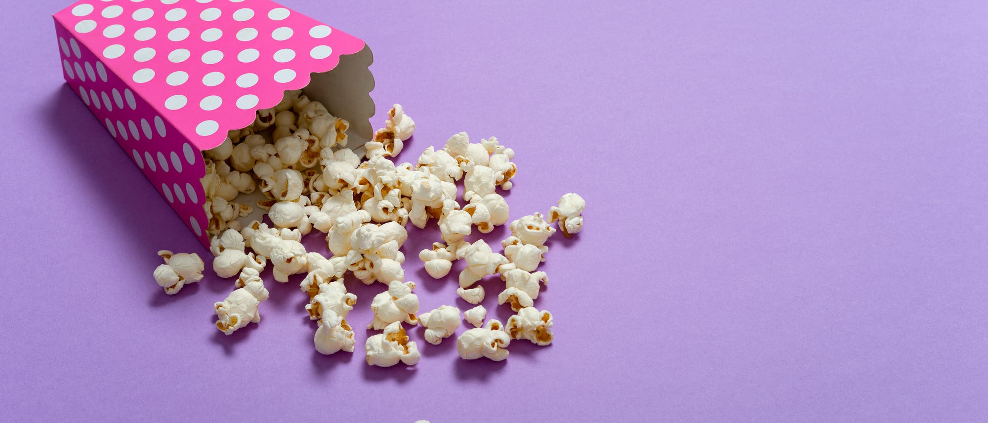 Eine pinkfarbene Popcorn-Schachtel mit weißen Punkten liegt auf einer lila Oberfläche. Popcorn ist aus der Schachtel herausgefallen und verteilt sich auf dem Untergrund. Eine pinkfarbene Popcorn-Schachtel mit weißen Punkten liegt auf einer lila Oberfläche. Popcorn ist aus der Schachtel herausgefallen und verteilt sich auf dem Untergrund.