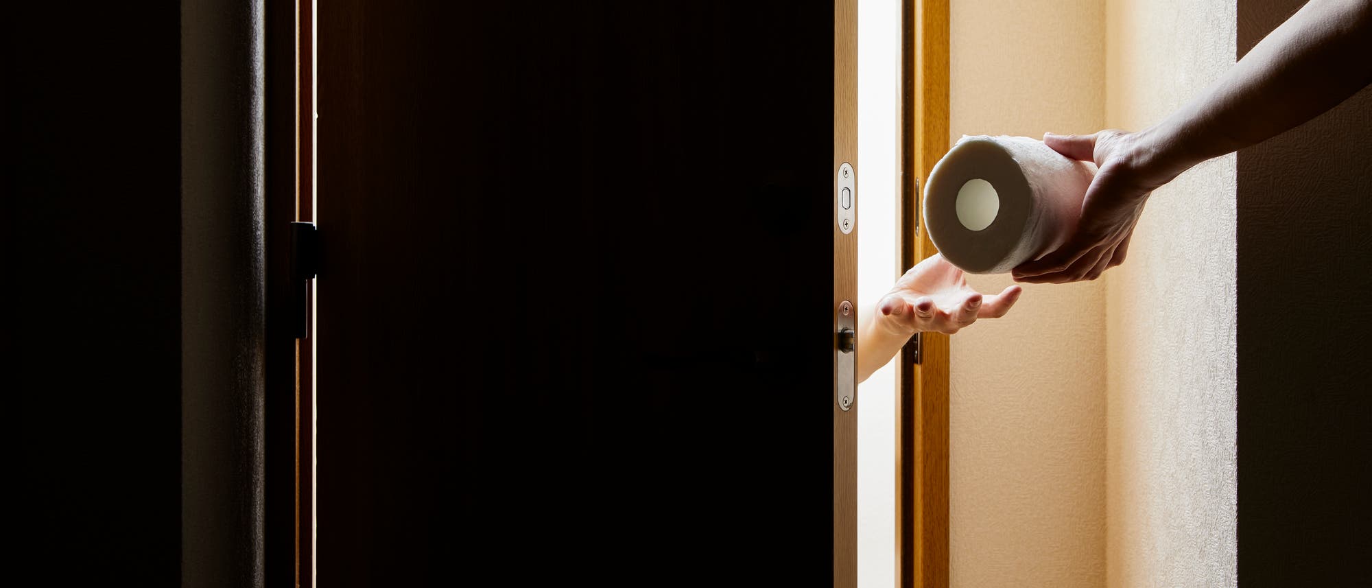 Eine Hand reicht eine Rolle Toilettenpapier durch einen leicht ge&ouml;ffneten T&uuml;rspalt in einen dunklen Raum. Das Bild zeigt den Moment des &Uuml;bergebens, wobei das Licht von au&szlig;en in den Raum f&auml;llt. Der Fokus liegt auf der Handlung des Gebens und Empfangens.