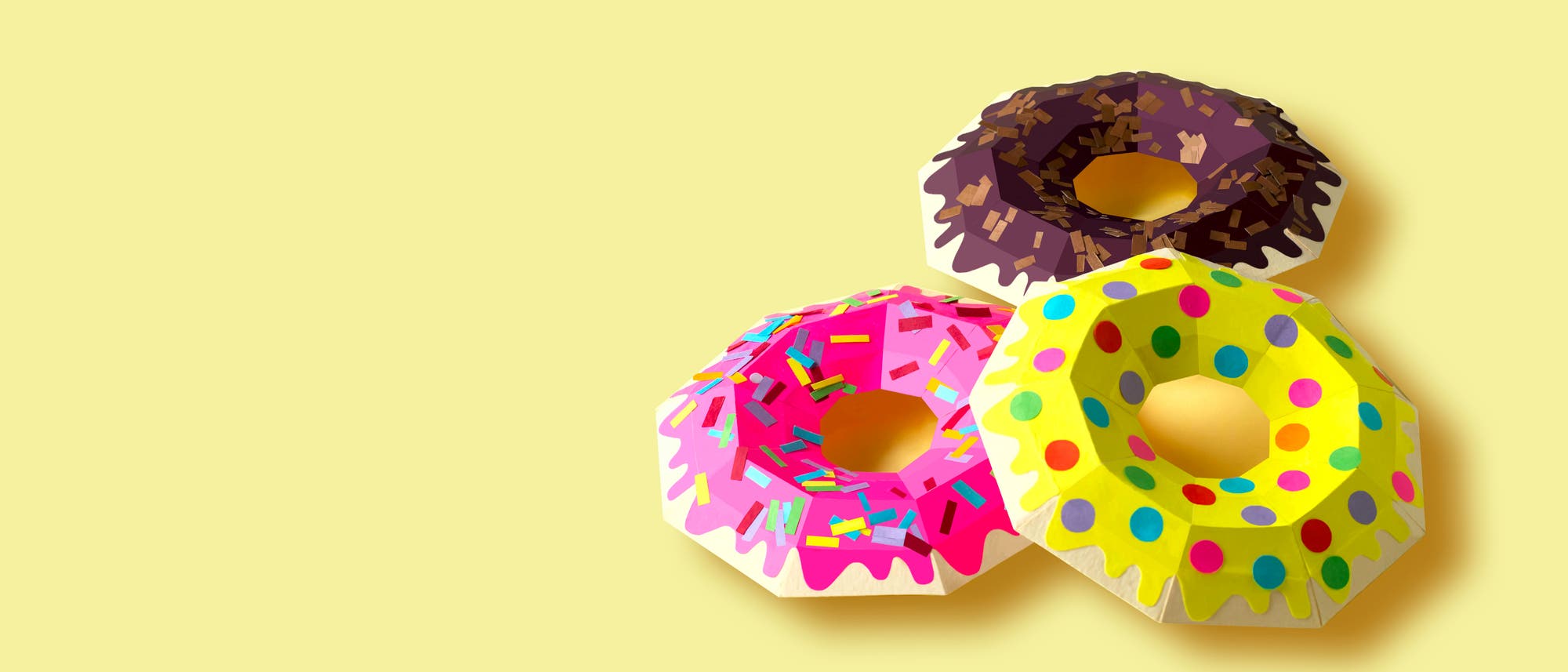 Drei bunte, geometrisch gestaltete Donuts aus Papier auf einem hellgelben Hintergrund. Jeder Donut hat ein einzigartiges Design: einer mit rosa Glasur und bunten Streuseln, ein anderer mit brauner Glasur und Schokoladenstückchen, und der dritte mit gelber Glasur und bunten Punkten. Die Komposition betont die kreative und künstlerische Darstellung von alltäglichen Objekten. Drei bunte, geometrisch gestaltete Donuts aus Papier auf einem hellgelben Hintergrund. Jeder Donut hat ein einzigartiges Design: einer mit rosa Glasur und bunten Streuseln, ein anderer mit brauner Glasur und Schokoladenstückchen, und der dritte mit gelber Glasur und bunten Punkten. Die Komposition betont die kreative und künstlerische Darstellung von alltäglichen Objekten.
