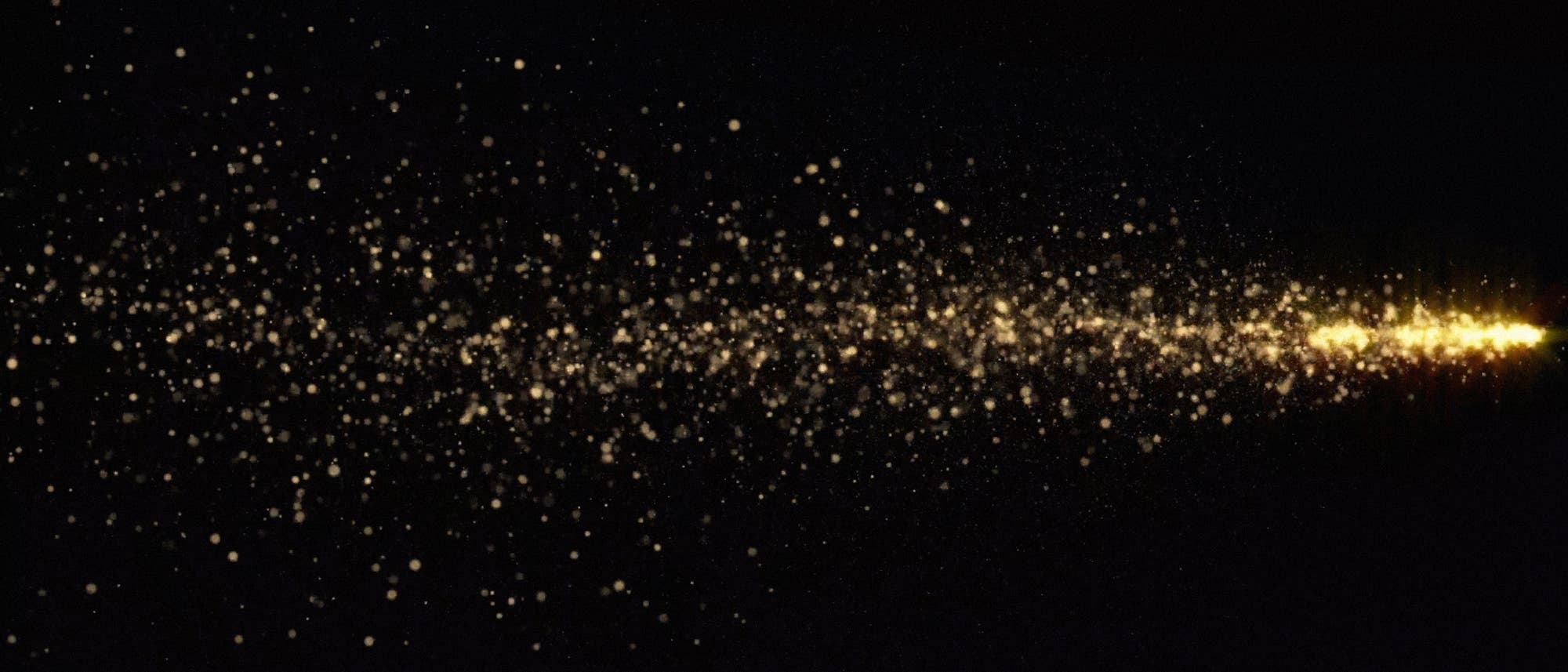 Ein Bild zeigt eine Ansammlung von goldenen Lichtpunkten, die sich von links nach rechts &uuml;ber einen schwarzen Hintergrund erstrecken. Die Punkte sind in der linken Bildh&auml;lfte dichter verteilt und werden nach rechts hin intensiver und heller, was den Eindruck eines leuchtenden Sternenstaubs oder eines kosmischen Ph&auml;nomens vermittelt.