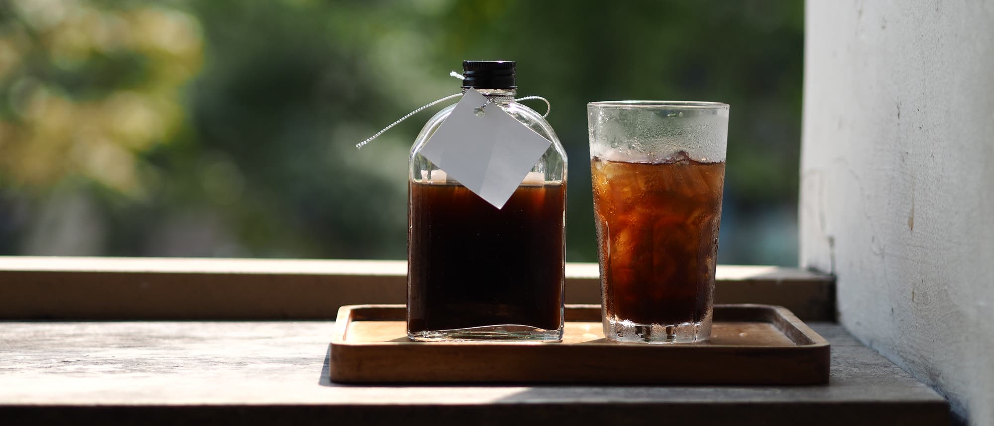 Eine Glasflasche mit dunklem Getränk steht neben einem Glas mit kaltem Kaffee und Eiswürfeln auf einem Holztablett in einer Fensteröffnung. Im Hintergrund sind unscharfe grüne Blätter zu sehen. Eine Glasflasche mit dunklem Getränk steht neben einem Glas mit kaltem Kaffee und Eiswürfeln auf einem Holztablett in einer Fensteröffnung. Im Hintergrund sind unscharfe grüne Blätter zu sehen.