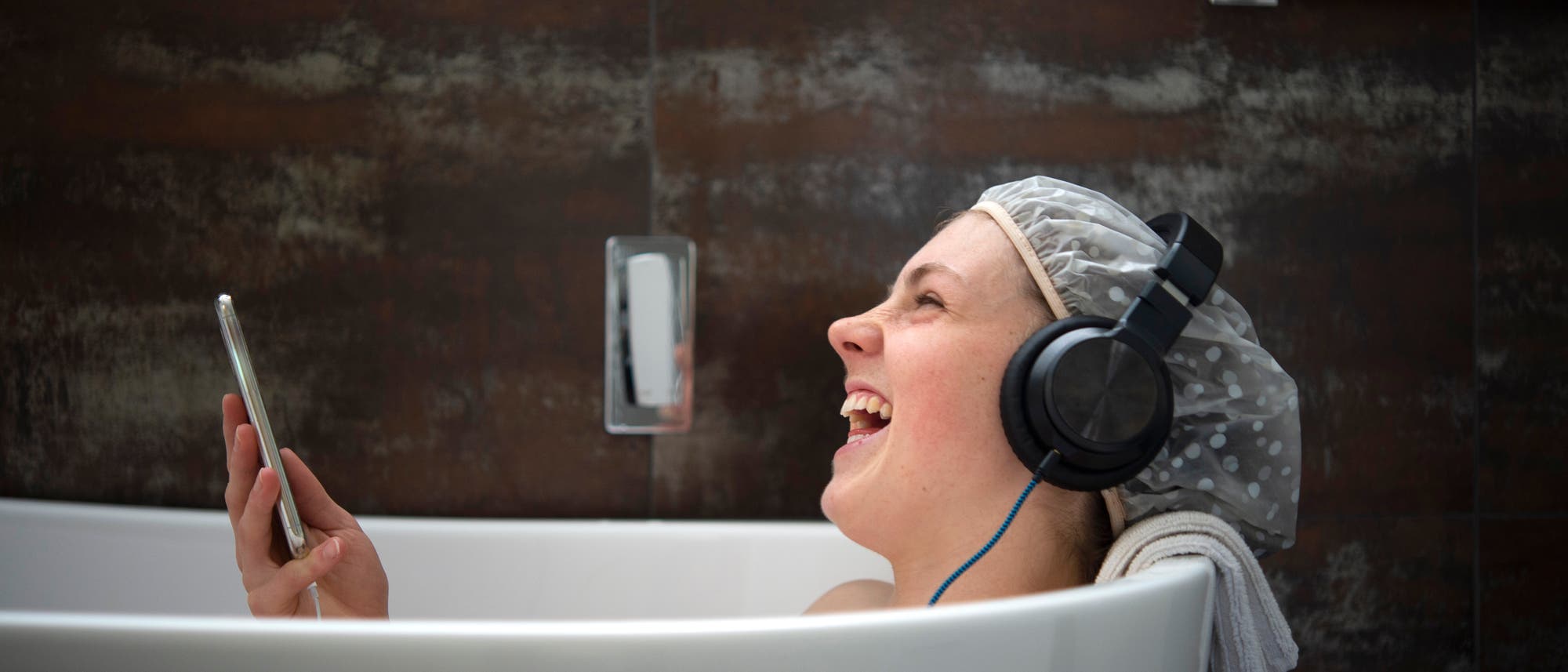 Eine junge Frau sitzt in einer Badewanne, trägt Kopfhörer und eine Duschhaube. Sie hält ein Smartphone in der Hand und lacht. Die Wand im Hintergrund ist dunkel und strukturiert. Die Szene vermittelt Spaß und Entspannung. Eine junge Frau sitzt in einer Badewanne, trägt Kopfhörer und eine Duschhaube. Sie hält ein Smartphone in der Hand und lacht. Die Wand im Hintergrund ist dunkel und strukturiert. Die Szene vermittelt Spaß und Entspannung.