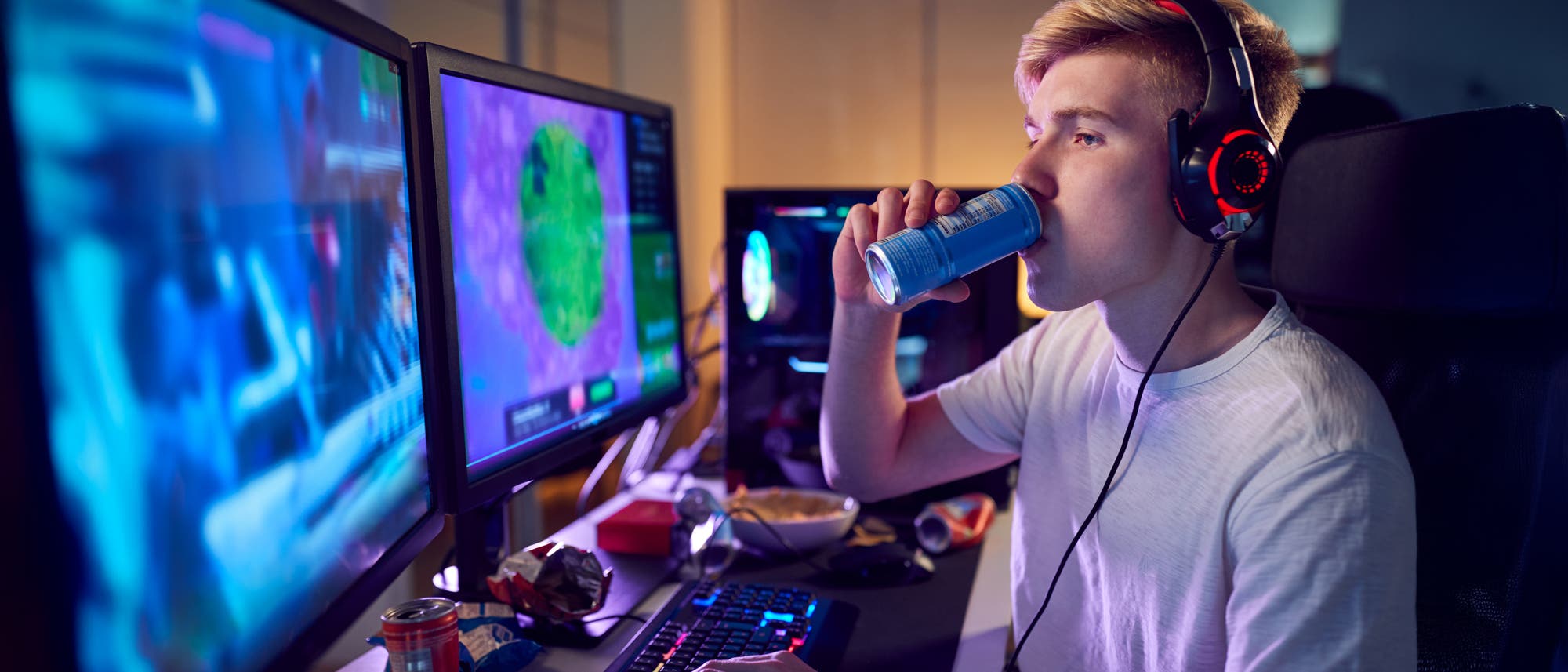 Ein junger Mensch sitzt an einem Schreibtisch mit mehreren Monitoren und spielt ein Videospiel. Er trägt Kopfhörer und trinkt aus einer Getränkedose. Auf dem Tisch befinden sich weitere Getränkedosen und Snackverpackungen. Die Monitore zeigen bunte Spielgrafiken. Die Szene ist in einem Raum mit gedämpfter Beleuchtung.