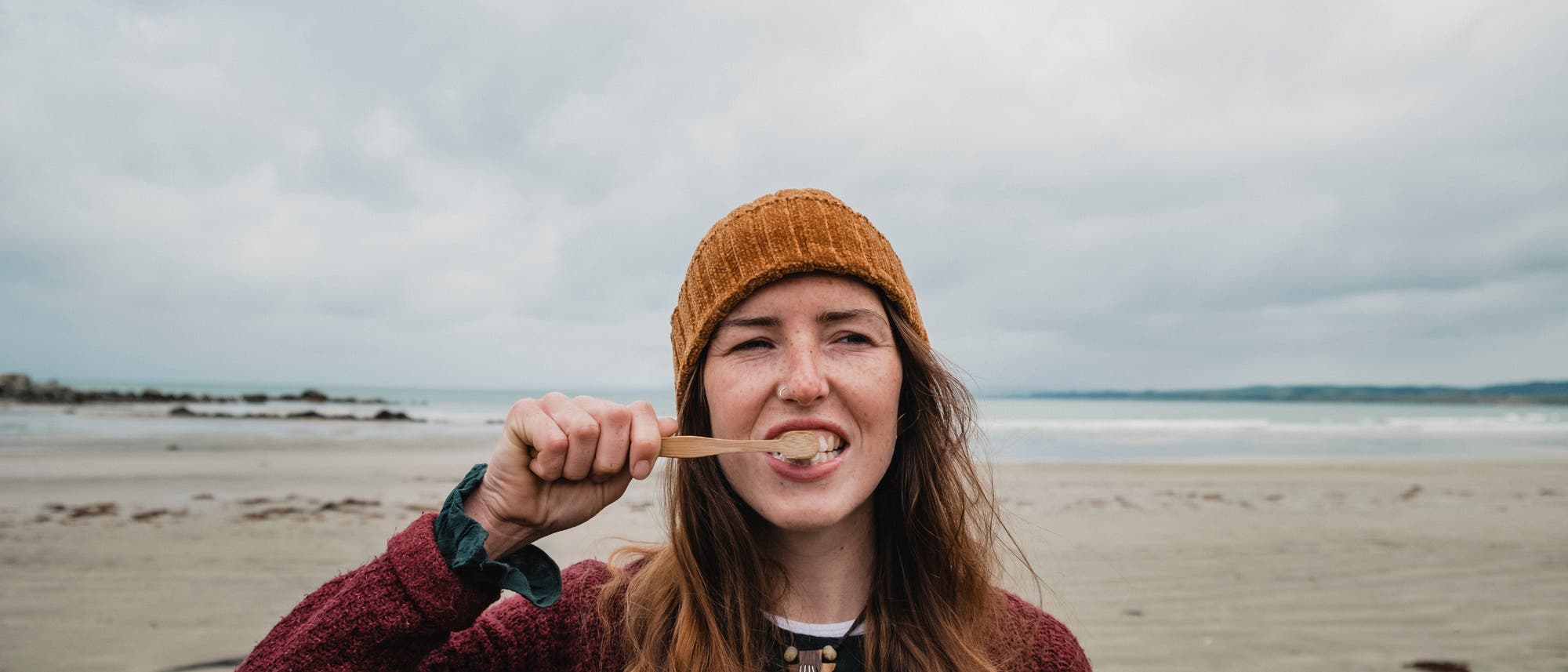 Eine Frau mit langen, rotbraunen Haaren und einer orangefarbenen Mütze putzt sich mit einer Bambuszahnbürste die Zähne. Sie trägt einen roten Pullover und steht an einem bewölkten Strand vor dem Meer. Im Hintergrund sind Wellen zu sehen.