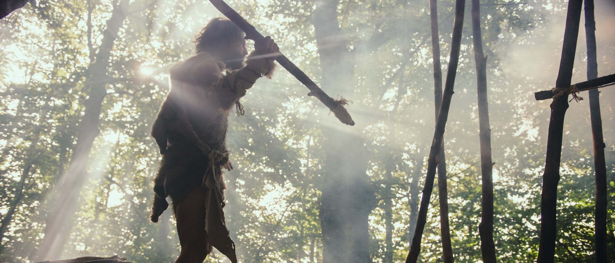 Eine Person in primitiver Kleidung steht in einem Wald, umgeben von Sonnenstrahlen, die durch die B&auml;ume scheinen. Die Person h&auml;lt einen langen Stock oder Speer und ist von rauchigem Dunst umgeben. Im Hintergrund sind B&auml;ume und ein provisorisches Ger&uuml;st aus Holzstangen zu sehen. Die Szene vermittelt eine pr&auml;historische Atmosph&auml;re.