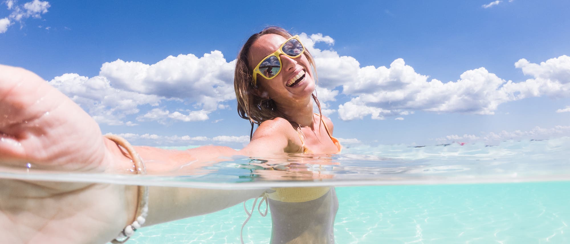 Eine Person macht ein Selfie im klaren, t&uuml;rkisfarbenen Wasser eines Strandes. Sie tr&auml;gt eine Sonnenbrille und l&auml;chelt in die Kamera. Der Himmel ist blau mit einigen wei&szlig;en Wolken. Die Aufnahme ist teilweise unter Wasser, was einen interessanten Effekt erzeugt.