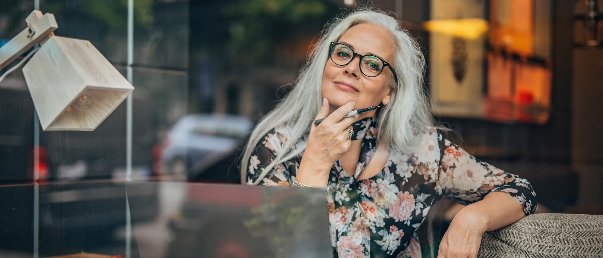 Eine Person sitzt in einem Café an einem Holztisch mit einem Laptop, einem Notizblock und einem Stift in der Hand. Die Person schaut nachdenklich aus dem Fenster. Im Hintergrund sind verschwommene Straßenszenen zu sehen.