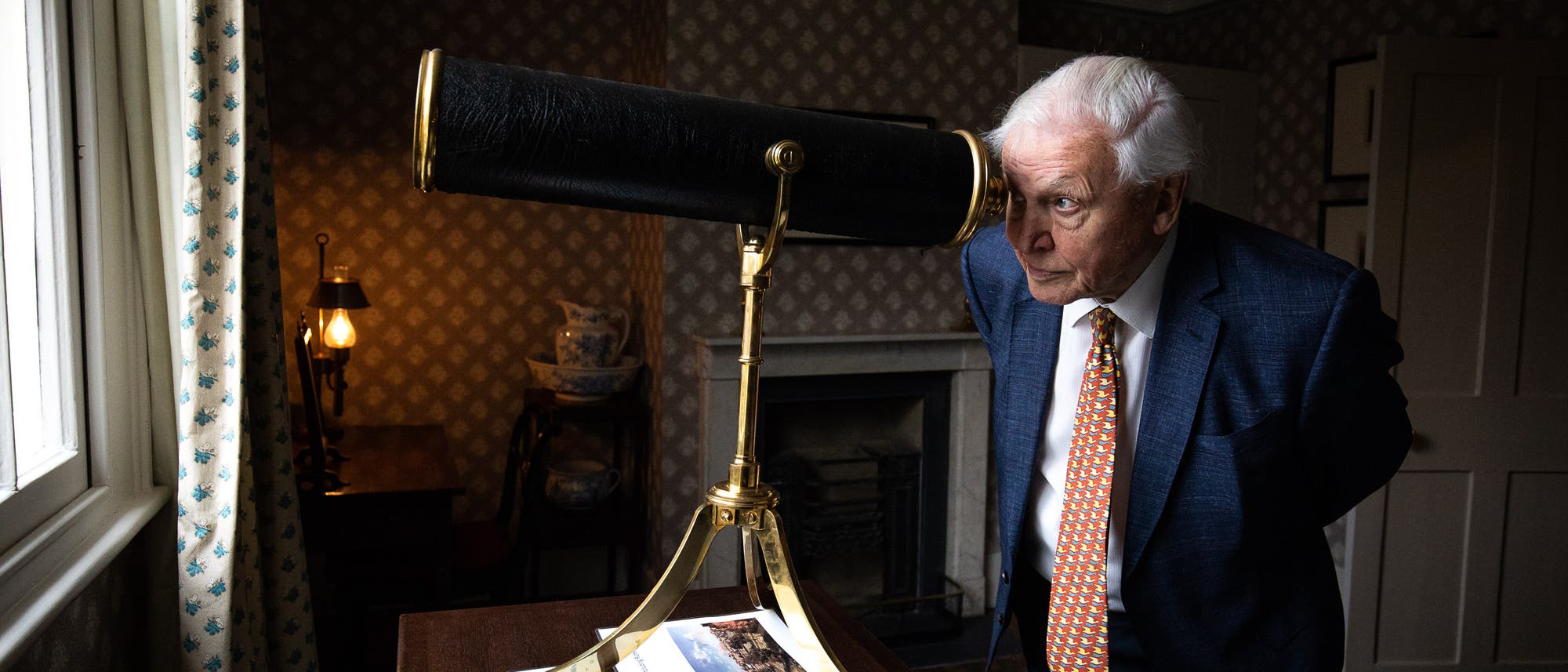 Dvid Attenborough schaut durch ein antikes Teleskop in einem traditionell eingerichteten Raum. Im Hintergrund sind eine Lampe und ein Kamin zu sehen. Die Szene vermittelt einen Eindruck von Neugier und Entdeckung.