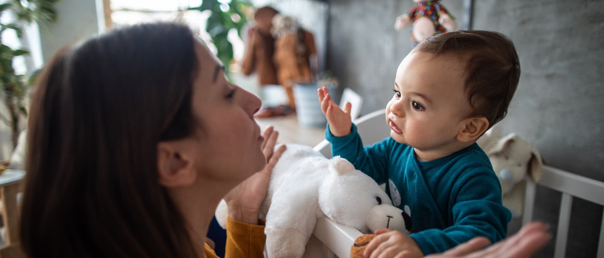 Eine Frau und ein Kleinkind interagieren in einem Kinderzimmer. Das Kind steht in einem Gitterbett und hält ein weißes Plüschtier, während es die Frau ansieht. Im Hintergrund sind Pflanzen und Regale mit Spielzeug und Büchern zu sehen. Die Szene vermittelt eine warme und familiäre Atmosphäre. Eine Frau und ein Kleinkind interagieren in einem Kinderzimmer. Das Kind steht in einem Gitterbett und hält ein weißes Plüschtier, während es die Frau ansieht. Im Hintergrund sind Pflanzen und Regale mit Spielzeug und Büchern zu sehen. Die Szene vermittelt eine warme und familiäre Atmosphäre.