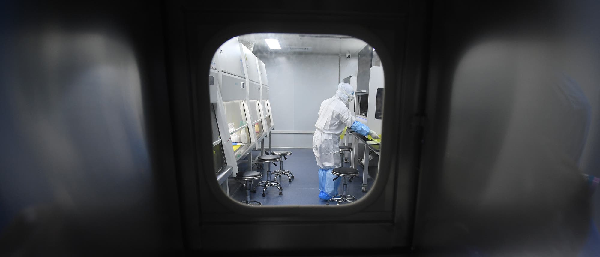 Eine Person in einem weißen Schutzanzug und blauen Handschuhen arbeitet in einem Zellkulturlabor mit mehreren Arbeitsstationen und Hockern. Die Szene wird durch ein Fenster von außen betrachtet. Eine Person in einem weißen Schutzanzug und blauen Handschuhen arbeitet in einem Zellkulturlabor mit mehreren Arbeitsstationen und Hockern. Die Szene wird durch ein Fenster von außen betrachtet.