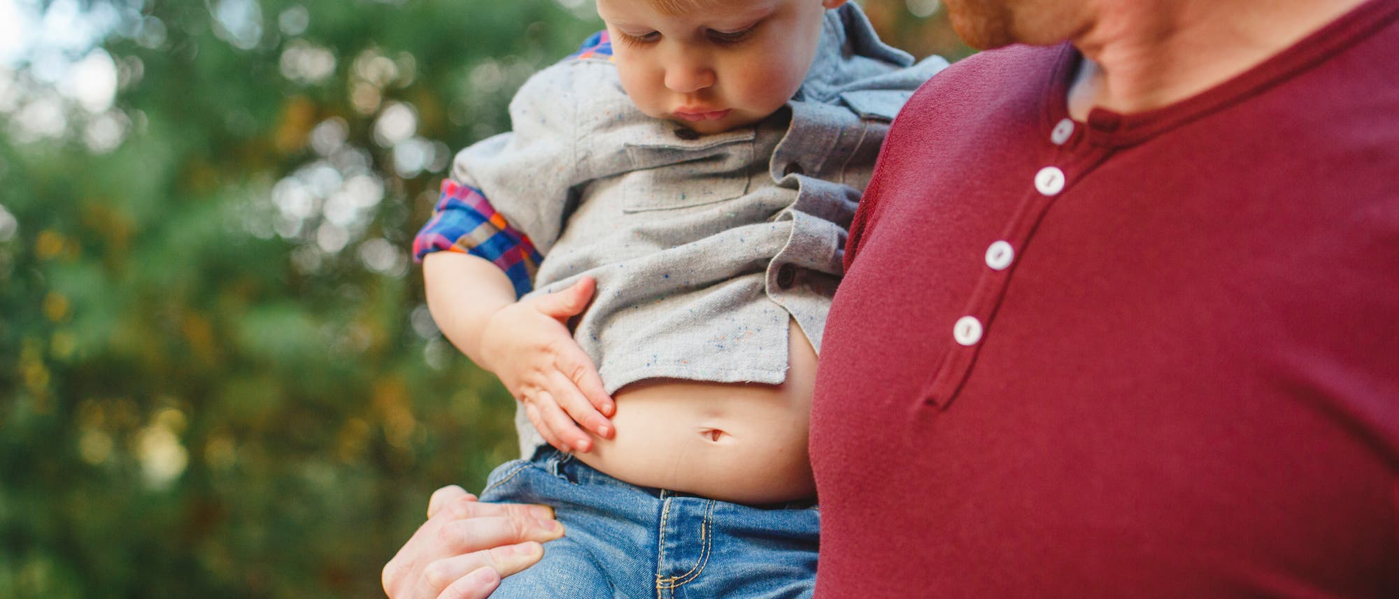 Ein Mann in einem roten Hemd hält ein kleines Kind, das neugierig auf seinen eigenen Bauchnabel schaut. Das Kind trägt ein graues Hemd mit karierten Details und blaue Jeans. Im Hintergrund sind verschwommene grüne Bäume zu sehen. Ein Mann in einem roten Hemd hält ein kleines Kind, das neugierig auf seinen eigenen Bauchnabel schaut. Das Kind trägt ein graues Hemd mit karierten Details und blaue Jeans. Im Hintergrund sind verschwommene grüne Bäume zu sehen.