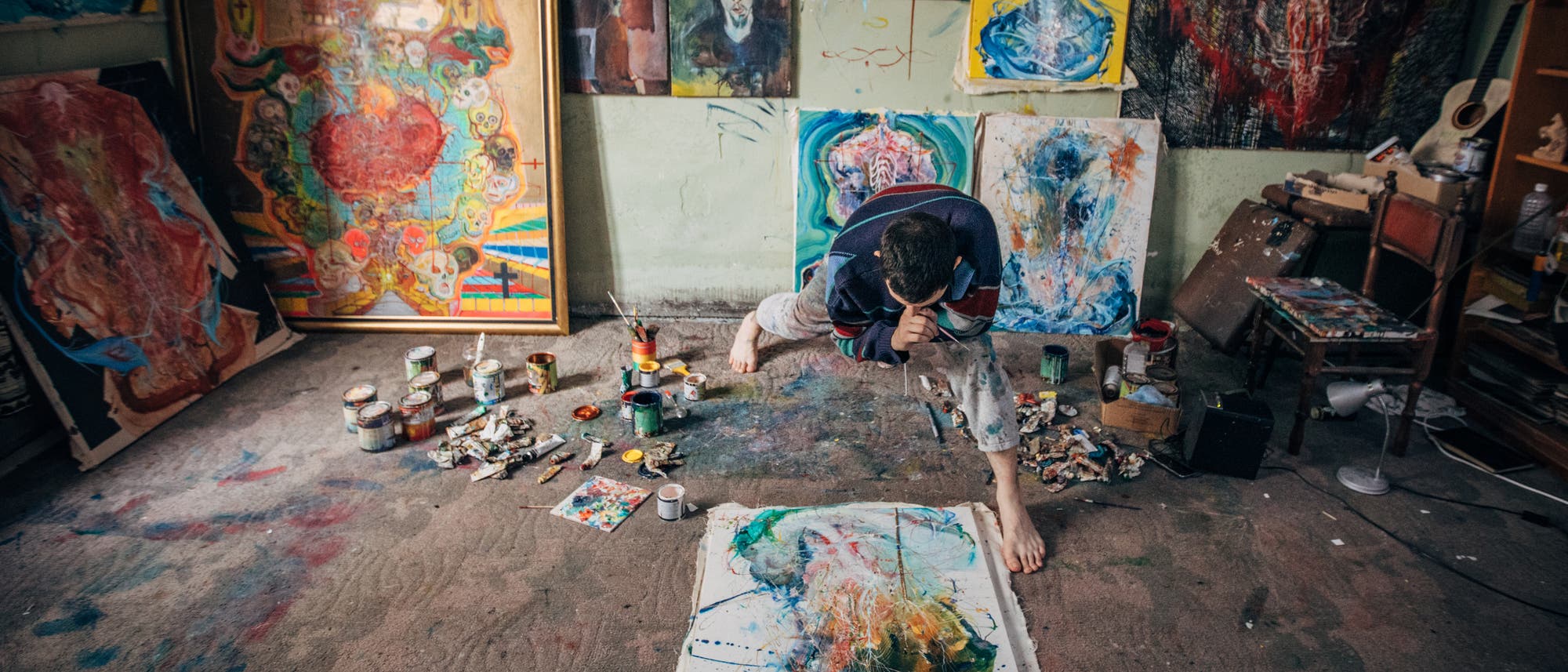 Ein Künstler arbeitet in einem Atelier, umgeben von farbenfrohen Gemälden und Kunstmaterialien. Auf dem Boden liegt eine Leinwand mit abstrakten Farbklecksen. Der Raum ist mit verschiedenen Kunstwerken an den Wänden dekoriert, die lebendige Farben und expressive Formen zeigen. Der Künstler ist in einer dynamischen Pose, während er an einem neuen Werk arbeitet. Farbdosen und Pinsel sind auf dem Boden verstreut, was auf einen kreativen und produktiven Arbeitsprozess hinweist. Ein Künstler arbeitet in einem Atelier, umgeben von farbenfrohen Gemälden und Kunstmaterialien. Auf dem Boden liegt eine Leinwand mit abstrakten Farbklecksen. Der Raum ist mit verschiedenen Kunstwerken an den Wänden dekoriert, die lebendige Farben und expressive Formen zeigen. Der Künstler ist in einer dynamischen Pose, während er an einem neuen Werk arbeitet. Farbdosen und Pinsel sind auf dem Boden verstreut, was auf einen kreativen und produktiven Arbeitsprozess hinweist.