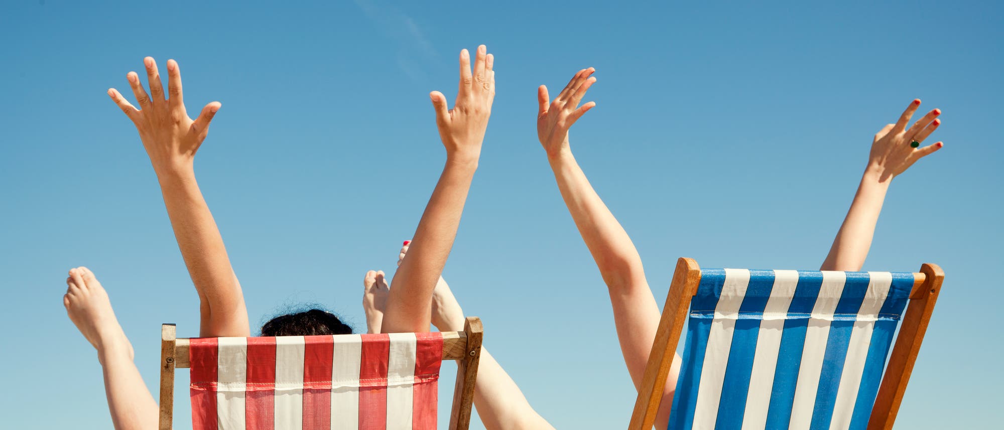 Zwei Personen sitzen in gestreiften Liegestühlen am Strand. Der linke Stuhl hat rote, der rechte blaue Streifen. Beide Personen heben ihre Arme und Beine in die Luft, während sie sich entspannen. Der Himmel ist klar und blau. Im Hintergrund ist das Meer zu sehen. Zwei Personen sitzen in gestreiften Liegestühlen am Strand. Der linke Stuhl hat rote, der rechte blaue Streifen. Beide Personen heben ihre Arme und Beine in die Luft, während sie sich entspannen. Der Himmel ist klar und blau. Im Hintergrund ist das Meer zu sehen.
