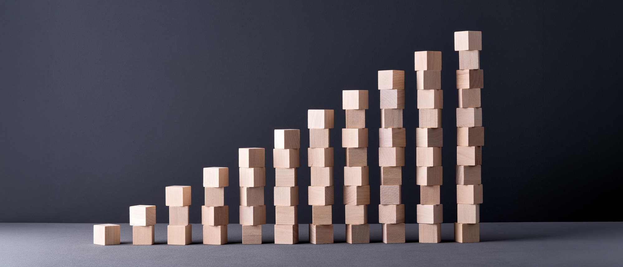 Eine Anordnung von Holzkl&ouml;tzen, die ein Balkendiagramm darstellen. Die Bausteine sind in aufsteigender Reihenfolge von links nach rechts gestapelt, was einen Anstieg oder Wachstum symbolisiert. Der Hintergrund ist dunkelgrau.
