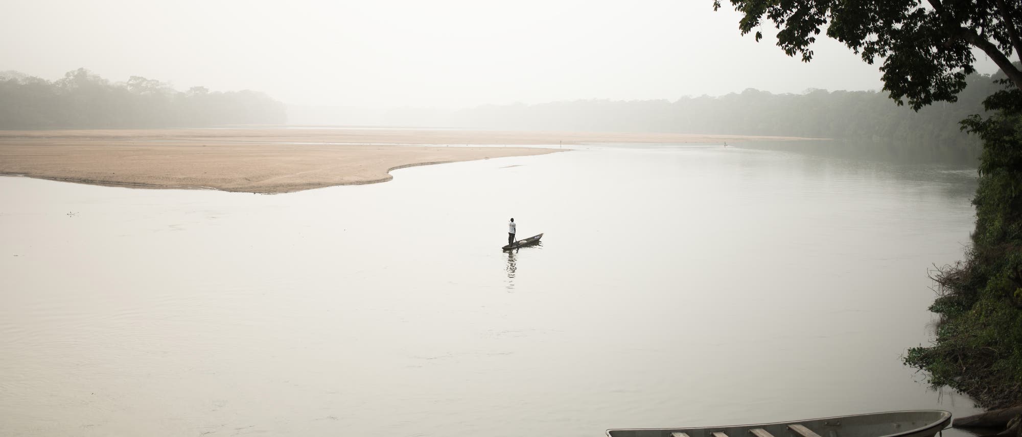 Eine ruhige Flusslandschaft bei Nebel. Ein einzelner Mensch steht in einem kleinen Boot in der Mitte des Flusses. Im Vordergrund sind zwei leere Boote am Ufer festgemacht. Im Hintergrund erstreckt sich eine sandige Landzunge, umgeben von dichter Vegetation. Ein großer Baum ragt von rechts ins Bild. Die Szene vermittelt eine friedliche und abgeschiedene Atmosphäre.
