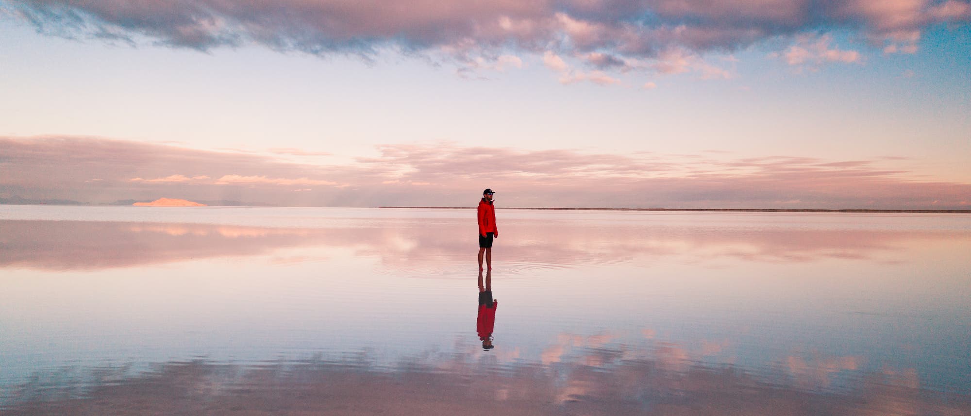 Eine Person in einer roten Jacke steht inmitten einer glatten Wasseroberfläche, auf der sich der Himmel mit rosa und blauen Wolken spiegelt. Der Horizont ist in der Ferne sichtbar, während die Wolken den Himmel dramatisch gestalten. Es handelt sich hier um die überfluteten Salzebenen "Bonneville Salt Flats" in Utah, USA.