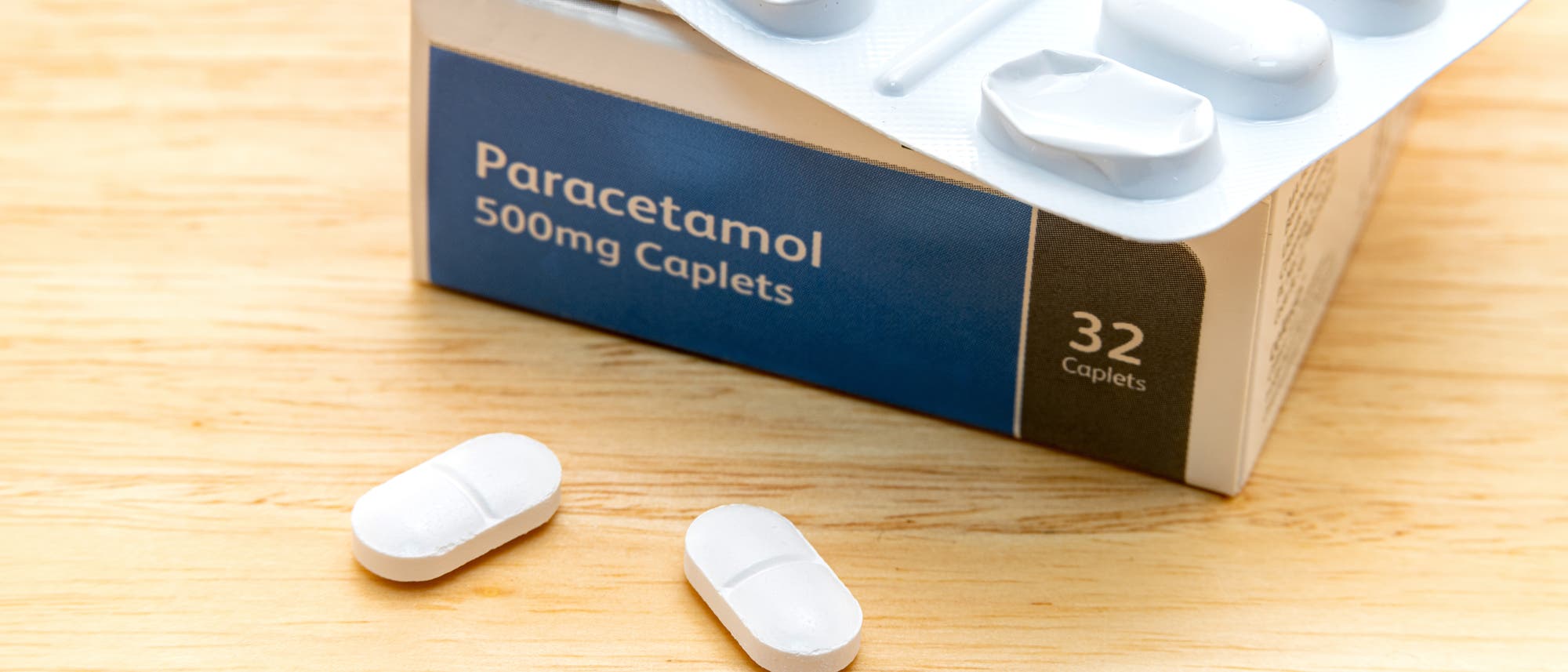 Eine Packung Paracetamol-Tabletten mit der Aufschrift „Paracetamol 500mg Caplets“ und „32 Caplets“ auf der Verpackung. Zwei weiße Tabletten liegen auf einer Holzoberfläche vor der geöffneten Blisterpackung.