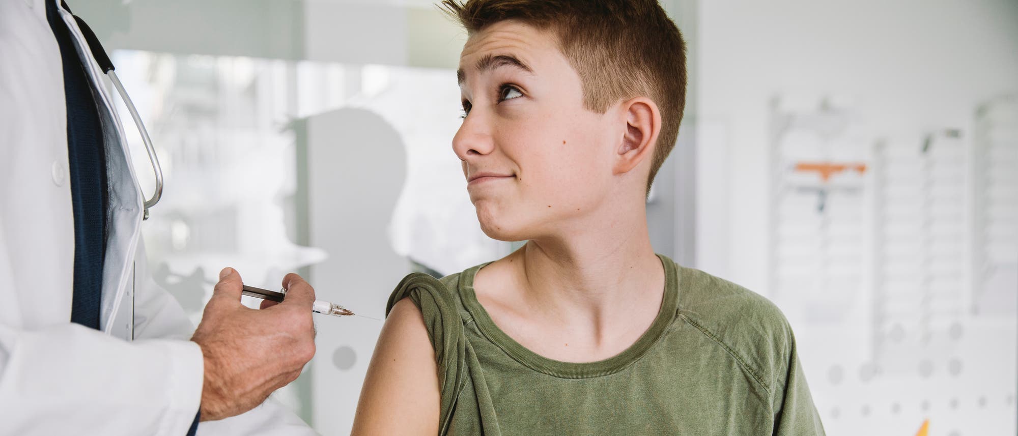 Ein Arzt in einem weißen Kittel verabreicht einem Jungen eine Impfung in den Oberarm. Der Junge trägt ein grünes T-Shirt und hält einen gelben Impfpass in der Hand. Die Szene spielt in einer medizinischen Umgebung mit unscharfem Hintergrund. Ein Arzt in einem weißen Kittel verabreicht einem Jungen eine Impfung in den Oberarm. Der Junge trägt ein grünes T-Shirt und hält einen gelben Impfpass in der Hand. Die Szene spielt in einer medizinischen Umgebung mit unscharfem Hintergrund.