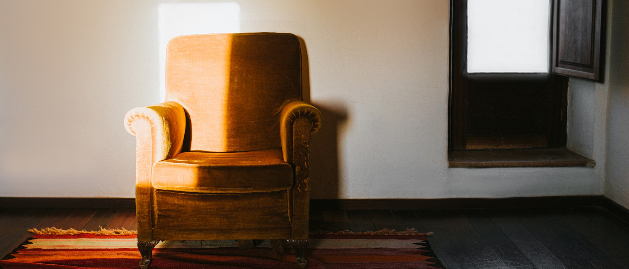 Ein Sessel mit goldbraunem Bezugsstoff steht vor einer hellen Wand auf einem gestreiften Teppich in einem sonst leeren Raum. Sonnenlicht fällt durch ein Fenster oder eine Tür auf den Sessel, wodurch ein heller Lichtstreifen auf einem Teil des Sessels entsteht. 