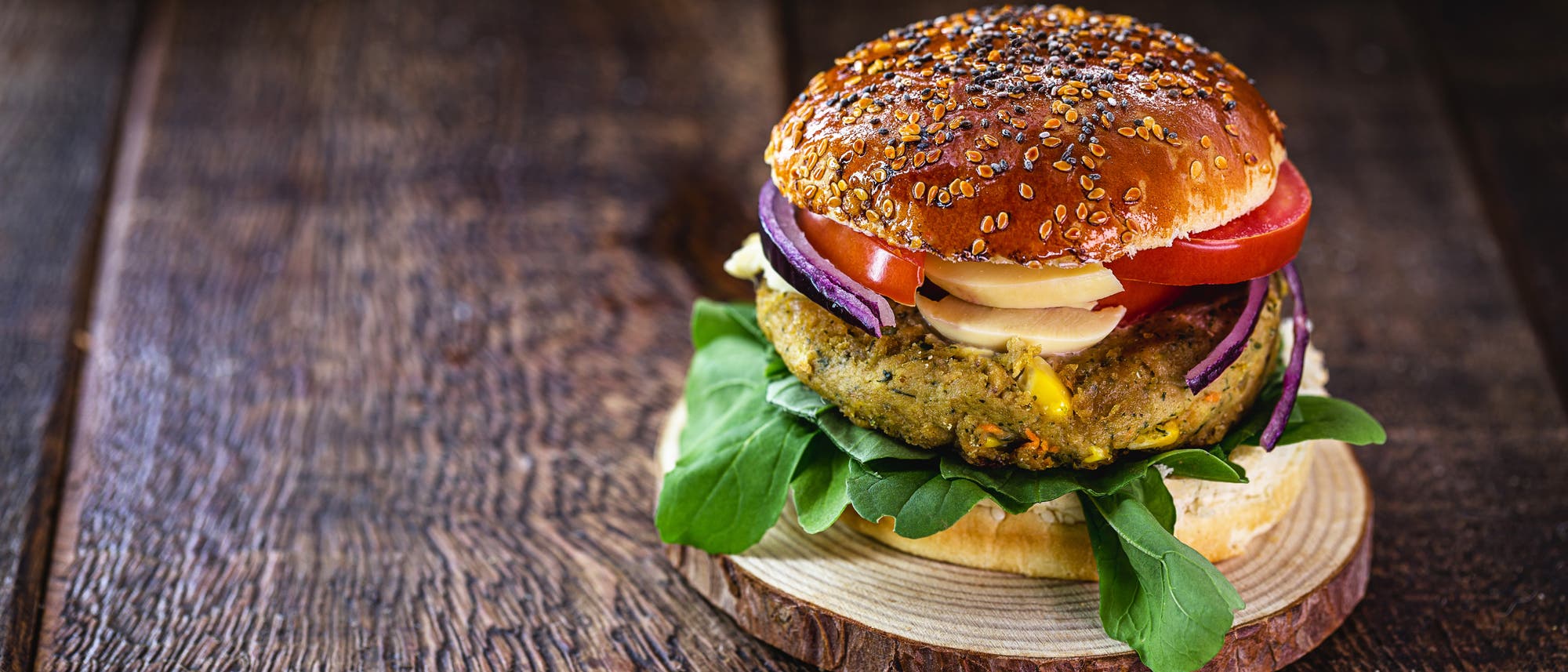 Ein vegetarischer Burger auf einem Holzbrett. Der Burger besteht aus einem Br&ouml;tchen mit Sesam, einem pflanzlichen Patty, frischen Tomatenscheiben, roten Zwiebelringen, K&auml;se und gr&uuml;nem Salat. Der Hintergrund ist eine rustikale Holzoberfl&auml;che.