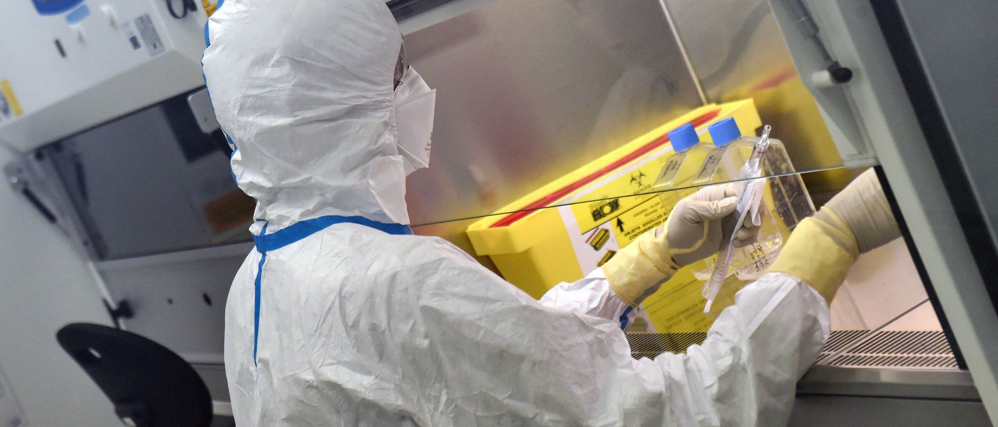 Eine Person in einem Schutzanzug arbeitet in einem Labor an einer Sicherheitswerkbank. Sie hält ein Laborgefäß und führt eine wissenschaftliche Untersuchung durch. Im Hintergrund sind Laborgeräte und ein gelber Behälter zu sehen. Die Szene vermittelt ein Gefühl von Präzision und Sicherheit in einem wissenschaftlichen Umfeld.