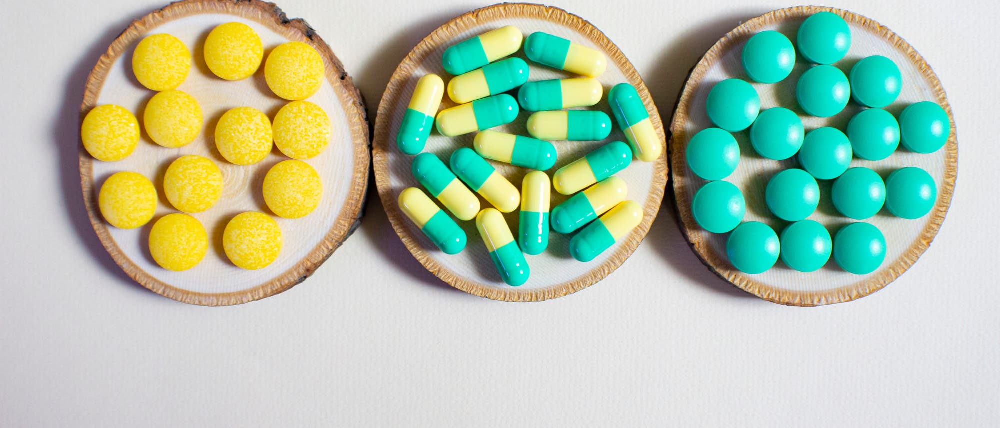 Drei runde Holzuntersetzer auf einer weißen Oberfläche, jeder mit einer anderen Art von Medikamenten: links gelbe Tabletten, in der Mitte gelb-türkise Kapseln, rechts türkisfarbene Tabletten. Drei runde Holzuntersetzer auf einer weißen Oberfläche, jeder mit einer anderen Art von Medikamenten: links gelbe Tabletten, in der Mitte gelb-türkise Kapseln, rechts türkisfarbene Tabletten.