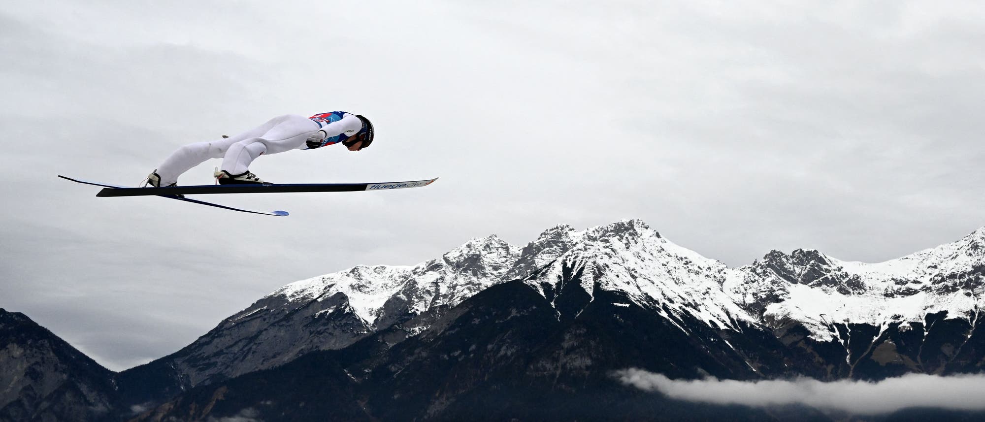 Ein Skispringer in weißem Anzug und Helm fliegt durch die Luft vor einer Kulisse aus schneebedeckten Bergen und bewölktem Himmel. Der Athlet ist in einer aerodynamischen Position, während er über die Landschaft gleitet. Ein Skispringer in weißem Anzug und Helm fliegt durch die Luft vor einer Kulisse aus schneebedeckten Bergen und bewölktem Himmel. Der Athlet ist in einer aerodynamischen Position, während er über die Landschaft gleitet.