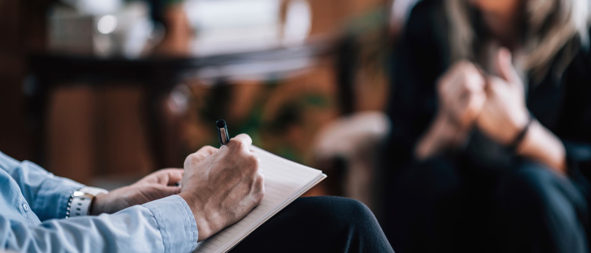 Eine Person sitzt mit einem Notizblock auf dem Schoß und schreibt mit einem Stift. Im Hintergrund ist eine weitere Person unscharf zu sehen, die möglicherweise in ein Gespräch vertieft ist. Die Szene deutet auf eine Beratung oder ein Interview hin. Der Raum wirkt gemütlich und professionell. Eine Person sitzt mit einem Notizblock auf dem Schoß und schreibt mit einem Stift. Im Hintergrund ist eine weitere Person unscharf zu sehen, die möglicherweise in ein Gespräch vertieft ist. Die Szene deutet auf eine Beratung oder ein Interview hin. Der Raum wirkt gemütlich und professionell.