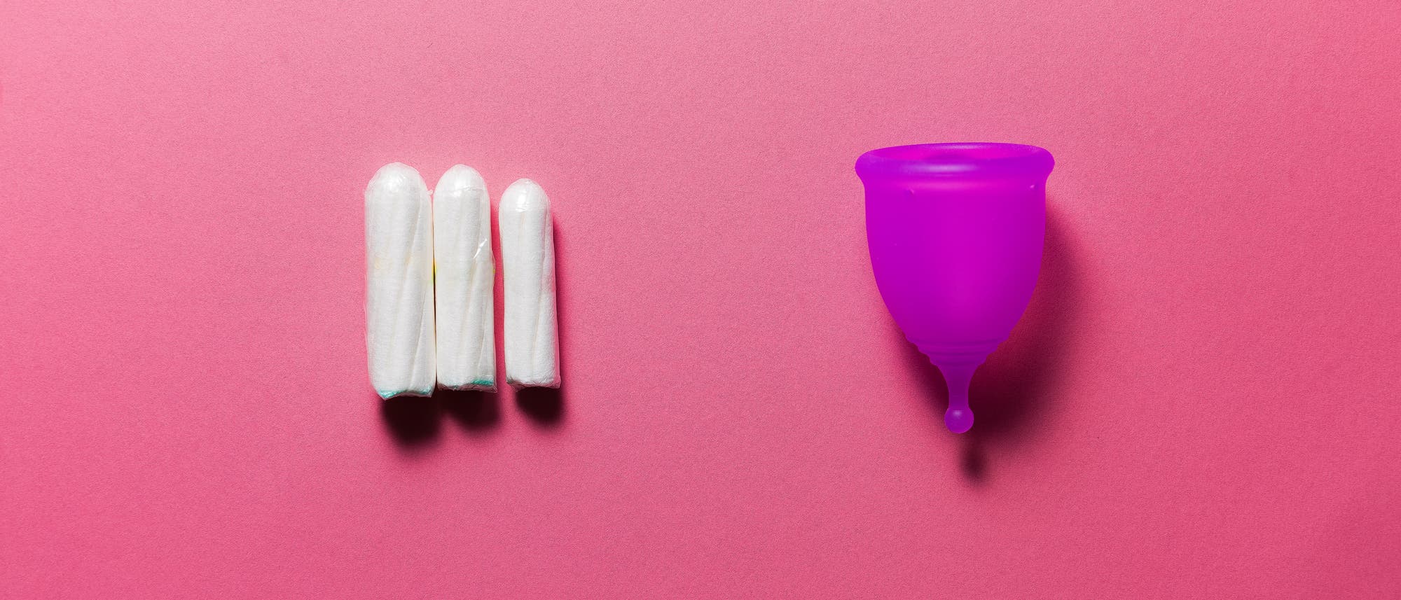 Drei weiße Tampons liegen nebeneinander auf einem rosa Hintergrund, links im Bild. Rechts daneben befindet sich eine lila Menstruationstasse. Das Bild vergleicht zwei Arten von Menstruationsprodukten. Drei weiße Tampons liegen nebeneinander auf einem rosa Hintergrund, links im Bild. Rechts daneben befindet sich eine lila Menstruationstasse. Das Bild vergleicht zwei Arten von Menstruationsprodukten.
