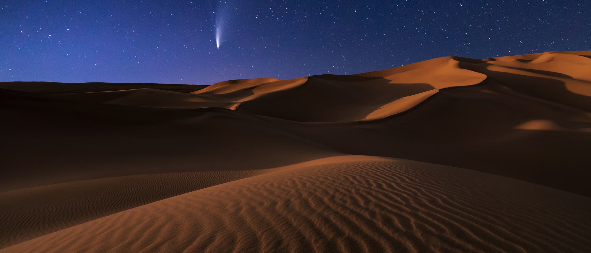Eine nächtliche Wüstenlandschaft mit sanften Sanddünen im Vordergrund, beleuchtet vom Mondlicht. Am klaren Sternenhimmel ist ein heller Komet mit einem langen Schweif zu sehen, der von links oben nach rechts unten verläuft. Die Szene vermittelt eine ruhige und majestätische Atmosphäre.