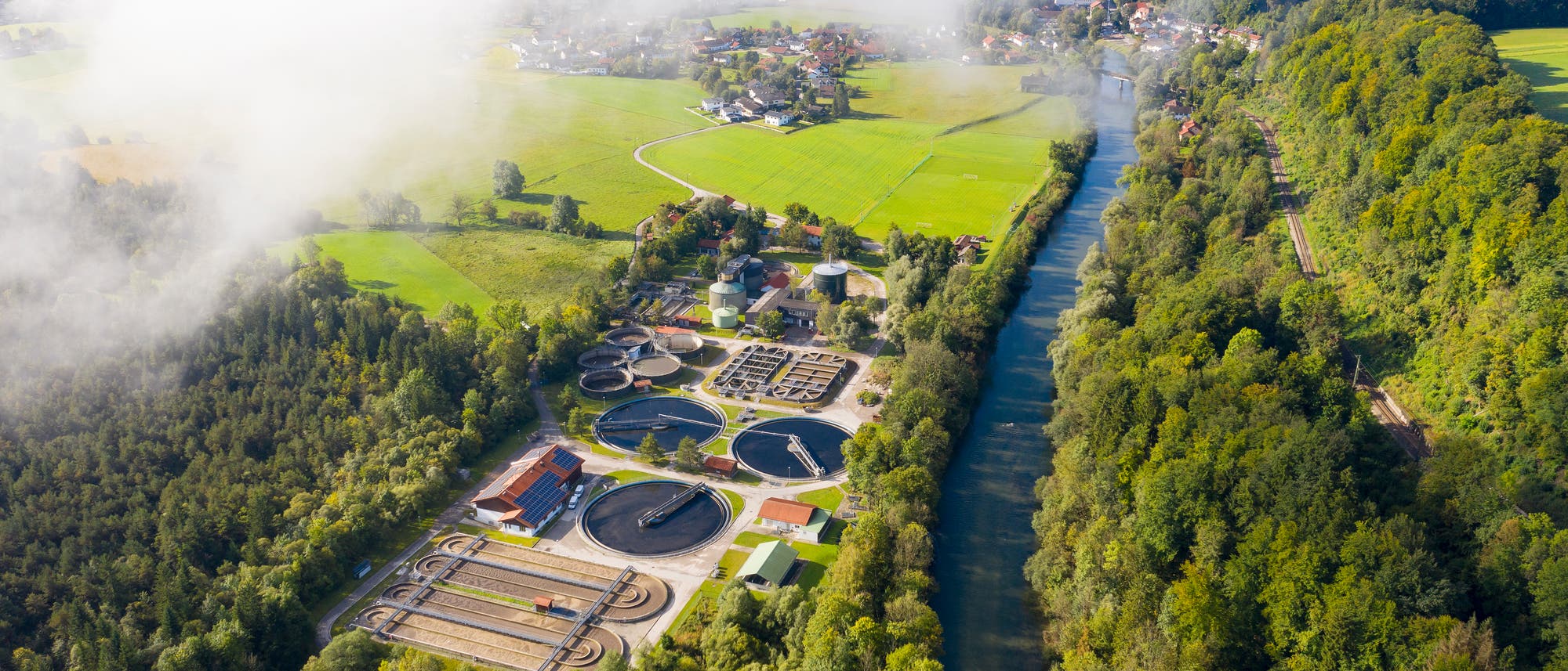 Luftaufnahme einer Kläranlage inmitten einer grünen Landschaft. Die Anlage besteht aus mehreren runden Becken und Gebäuden, die von Bäumen umgeben sind. Ein Fluss fließt rechts parallel zur Anlage, während im Hintergrund Felder und eine kleine Siedlung zu sehen sind. 