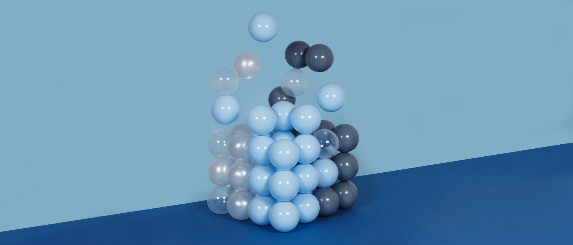 Ein Stapel aus hellblauen, dunkelblauen und durchsichtigen Kugeln, die an einer Wand aufeinandergestapelt sind. Die Wand ist in einem hellen Blauton gehalten, während der Boden dunkelblau ist. Einige Kugeln scheinen sich vom Stapel zu lösen und schweben nach oben.