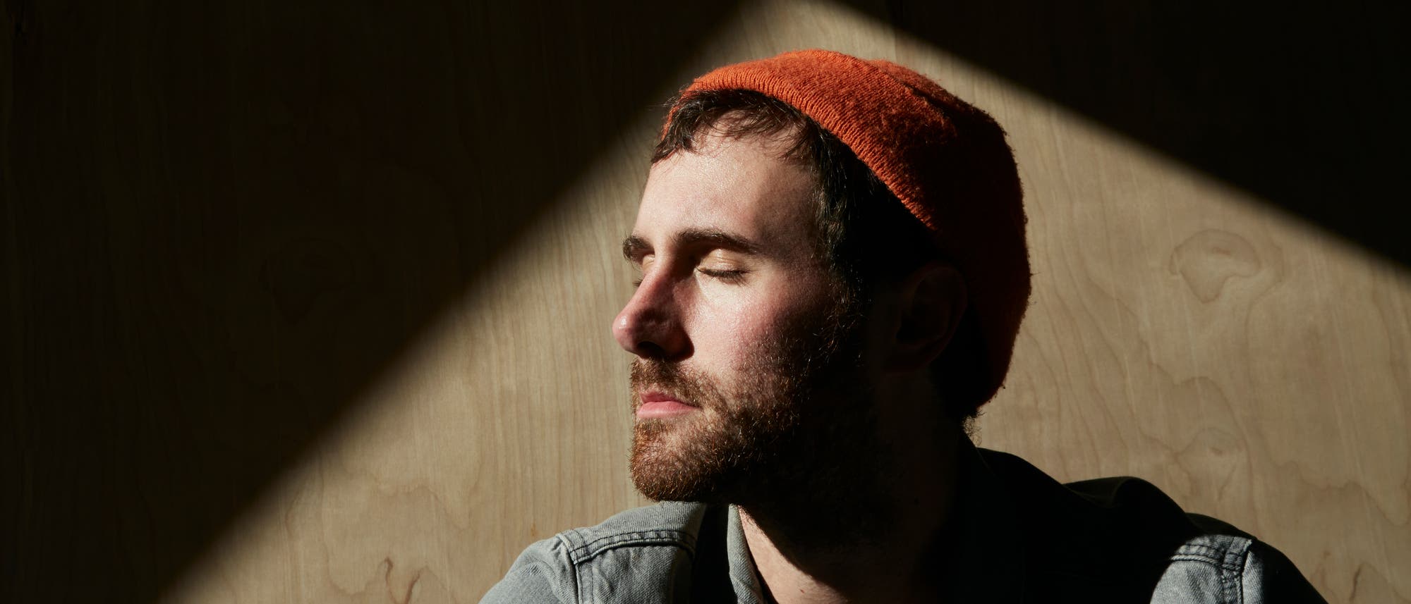 Ein Mann mit Jeanshemd und einer roten Strickmütze schaut seitlich und genießt mit geschlossenen Augen das Sonnenlicht, das auf sein Gesicht scheint. 