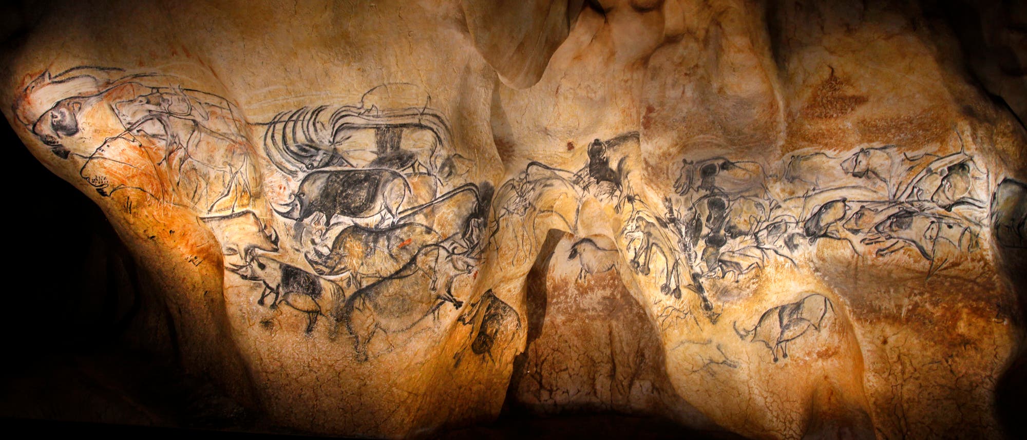 Eine H&ouml;hlenwand mit pr&auml;historischen Felsmalereien, die verschiedene Tiere wie Pferde, Rinder und Hirsche darstellen. Die Zeichnungen sind in dunklen Linien auf dem unebenen, nat&uuml;rlichen Felsuntergrund ausgef&uuml;hrt. Die Szene ist in warmes Licht getaucht, das die Details der Kunstwerke hervorhebt. Diese Malereien sind ein Beispiel f&uuml;r fr&uuml;he menschliche Kunst und zeigen die Bedeutung von Tieren in der pr&auml;historischen Kultur. Es handelt sich allerdings um eine detailgetreue Rekonstruktion.