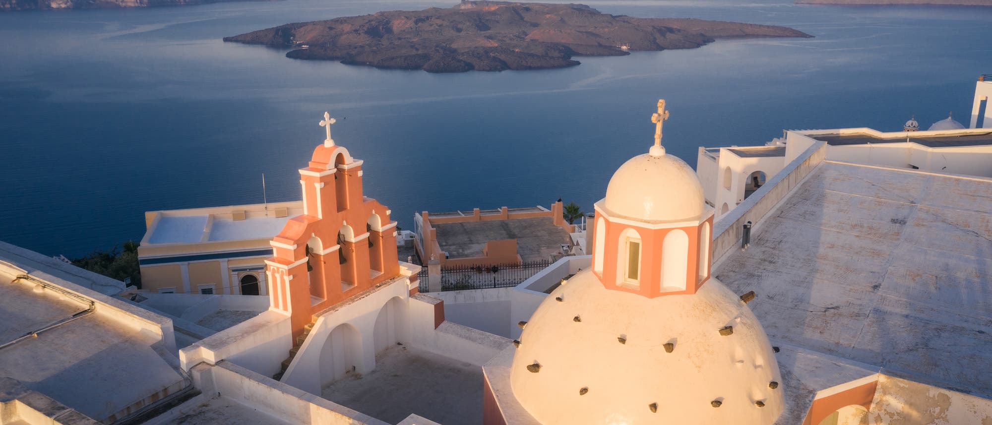 Eine malerische Ansicht von Santorini, Griechenland, mit einer traditionellen Kirche im Vordergrund, die eine cremefarbene Kuppel und orangefarbene Glockentürme aufweist. Im Hintergrund erstreckt sich die tiefblaue Ägäis mit einer vulkanischen Insel in der Mitte. Die Architektur und die natürliche Schönheit der Umgebung werden bei Sonnenuntergang beleuchtet.