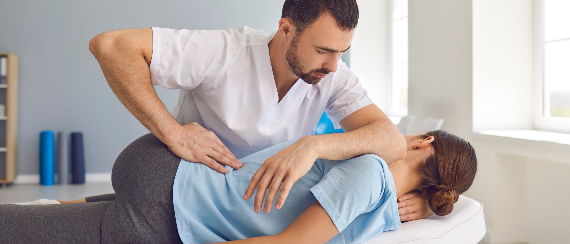 Ein Therapeut führt eine manuelle Therapie an einer liegenden Patientin durch. Die Patientin liegt auf einer Behandlungsliege in einem hellen Raum. Der Therapeut trägt ein weißes Oberteil und konzentriert sich auf die Behandlung. Im Hintergrund sind Fenster und einige Fitnessrollen zu sehen. Ein Therapeut führt eine manuelle Therapie an einer liegenden Patientin durch. Die Patientin liegt auf einer Behandlungsliege in einem hellen Raum. Der Therapeut trägt ein weißes Oberteil und konzentriert sich auf die Behandlung. Im Hintergrund sind Fenster und einige Fitnessrollen zu sehen.