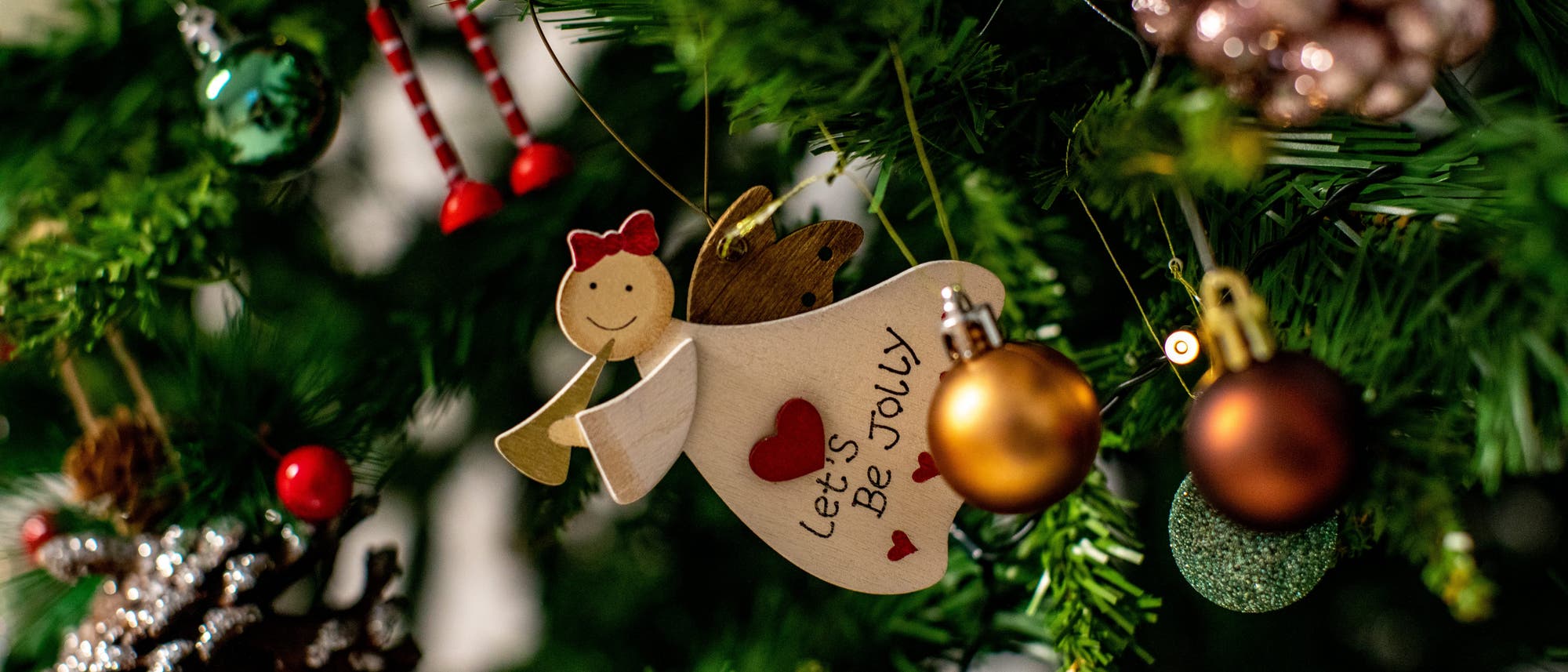 Ein geschm&uuml;ckter Weihnachtsbaum mit verschiedenen Dekorationen, darunter ein h&ouml;lzerner Engel mit der Aufschrift &bdquo;Let's Be Jolly&ldquo;, rote und goldene Kugeln, eine Tannenzapfenverzierung und ein Paar rot-wei&szlig; gestreifte Beine. Der Baum ist mit gr&uuml;nen Nadeln und Lichtern geschm&uuml;ckt, die eine festliche Atmosph&auml;re schaffen.