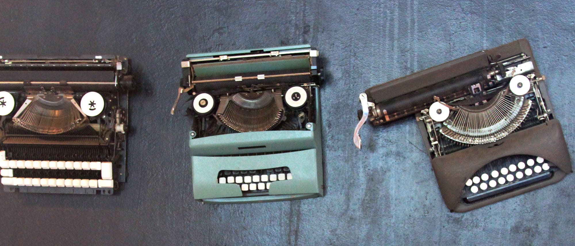 Drei antike Schreibmaschinen sind an einer blauen Wand befestigt. Jede Schreibmaschine ist so positioniert, dass sie wie ein Gesicht aussieht, wobei die Tastenreihen wie Z&auml;hne wirken. Die Schreibmaschinen sind in verschiedenen Winkeln angebracht, was den Eindruck von unterschiedlichen Gesichtsausdr&uuml;cken verst&auml;rkt.