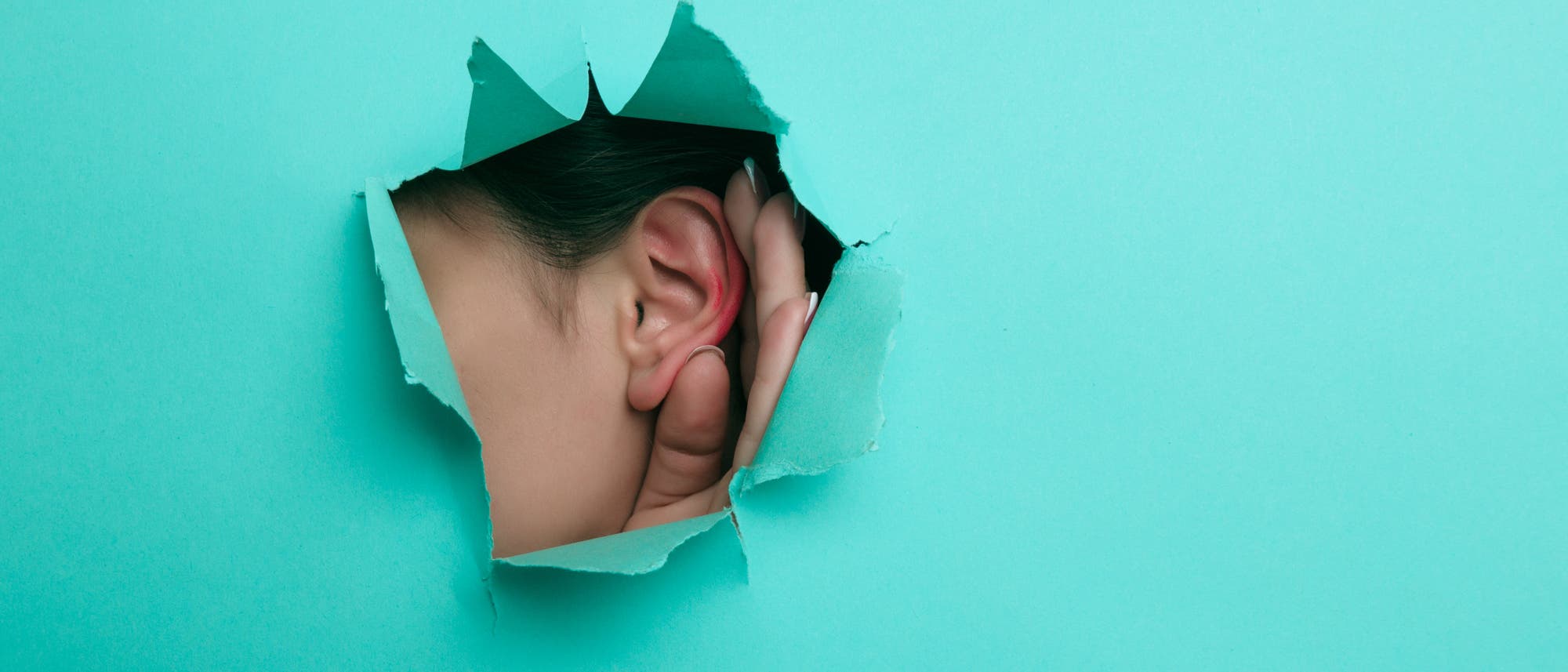 Eine Person hält ihre Hand an ihr Ohr und lauscht durch ein Loch in einer türkisfarbenen Papierwand. Das Bild symbolisiert das Konzept des Zuhörens oder Lauschens. Eine Person hält ihre Hand an ihr Ohr und lauscht durch ein Loch in einer türkisfarbenen Papierwand. Das Bild symbolisiert das Konzept des Zuhörens oder Lauschens.