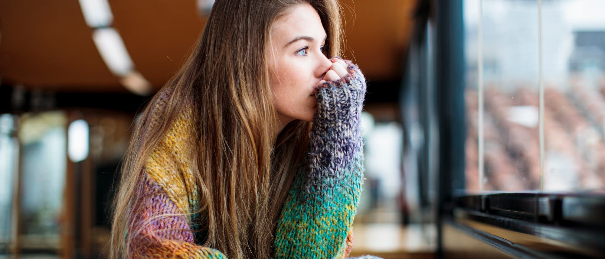 Eine Person sitzt an einem Tisch in einem Caf&eacute; und schaut nachdenklich aus dem Fenster. Sie tr&auml;gt einen bunten Strickpullover. Im Hintergrund sind verschwommene Lichter und moderne Architektur zu sehen. Die Szene vermittelt eine ruhige und nachdenkliche Stimmung.
