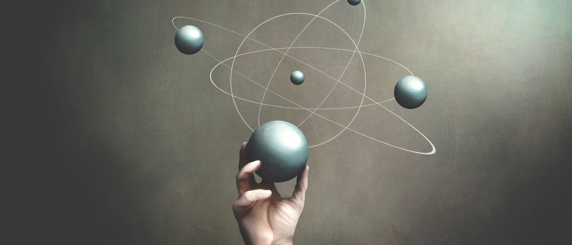 Eine Hand hält eine große, silberne Kugel, umgeben von kleineren Kugeln und weißen Linien, die an ein Atommodell erinnern. Der Hintergrund ist schlicht und dunkel, wodurch die Kugeln hervorgehoben werden. Die Szene vermittelt ein Gefühl von Wissenschaft und Technologie.