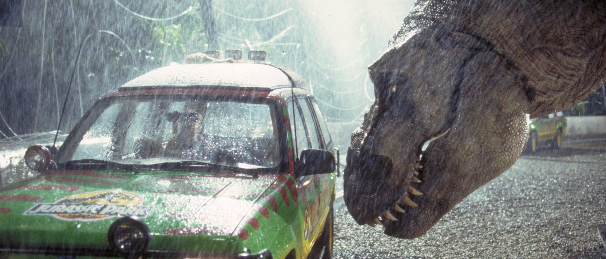Ein Dinosaurier beugt sich bedrohlich über ein buntes Fahrzeug mit dem Logo „Jurassic Park“ im Regen. Das Auto steht auf einem kiesigen Weg, umgeben von hohen Zäunen. Die Szene vermittelt Spannung und Gefahr. Ein Dinosaurier beugt sich bedrohlich über ein buntes Fahrzeug mit dem Logo „Jurassic Park“ im Regen. Das Auto steht auf einem kiesigen Weg, umgeben von hohen Zäunen. Die Szene vermittelt Spannung und Gefahr.