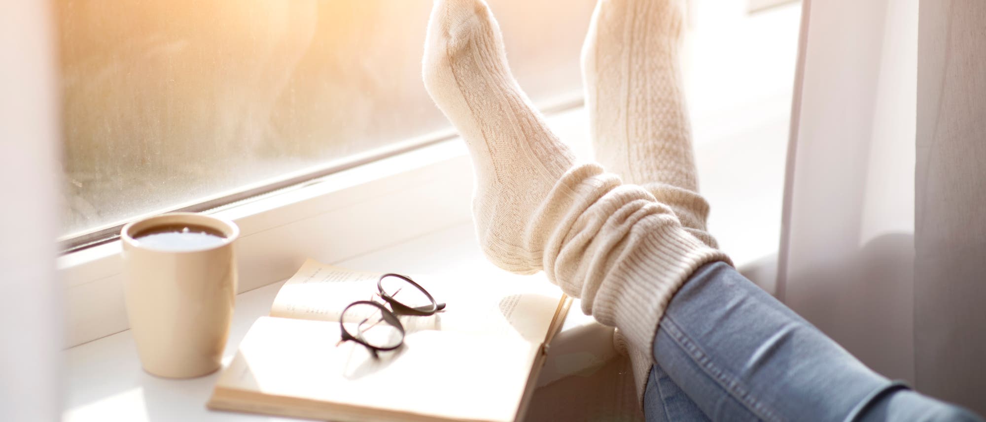 Fensterbank mit Buch und Brille darauf, einer Tasse Kaffee und hochgelegten Füßen, durchs Fenster scheint die Sonne