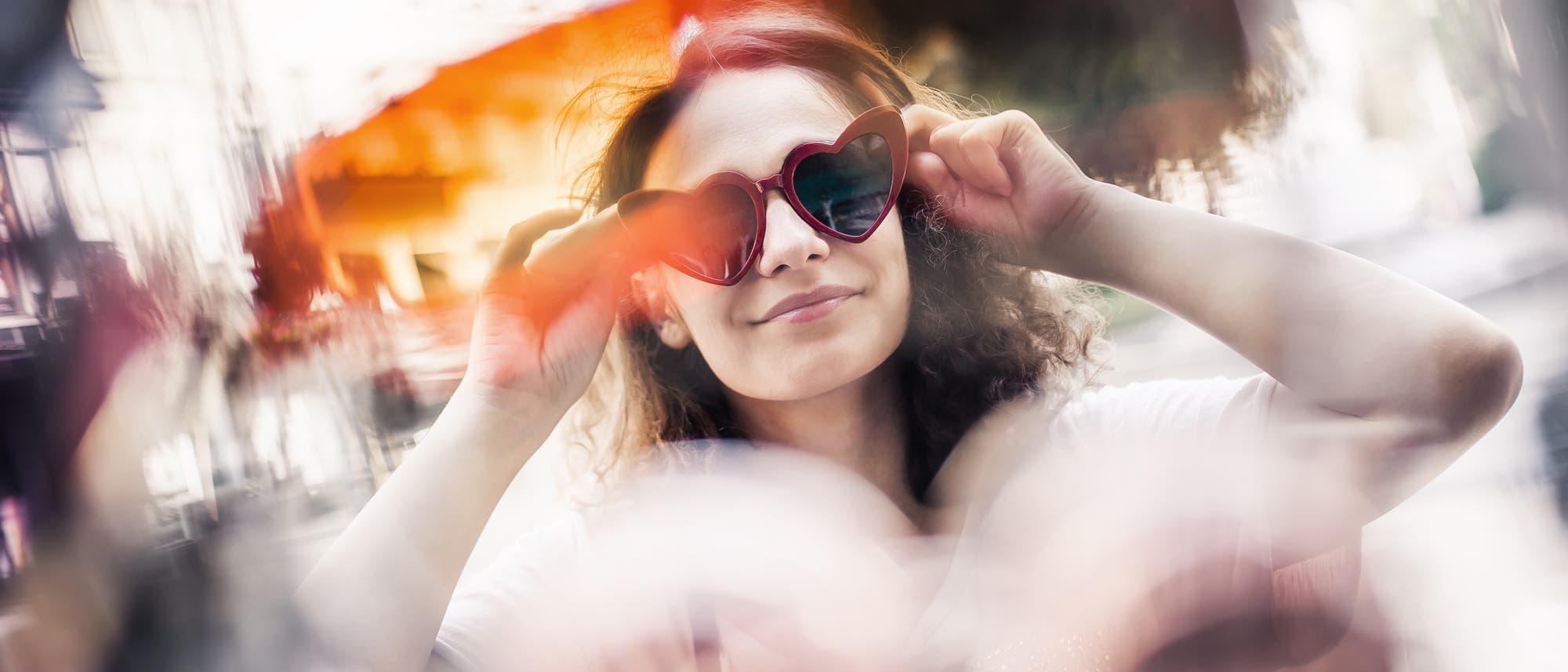 Eine Frau mit Locken trägt eine herzförmige Sonnenbrille und lächelt leicht. Die Umgebung ist verschwommen, was einen künstlerischen Effekt erzeugt. Die Farben sind warm und lebendig. Die Szene vermittelt ein Gefühl von Freude und Unbeschwertheit. Eine Frau mit Locken trägt eine herzförmige Sonnenbrille und lächelt leicht. Die Umgebung ist verschwommen, was einen künstlerischen Effekt erzeugt. Die Farben sind warm und lebendig. Die Szene vermittelt ein Gefühl von Freude und Unbeschwertheit.