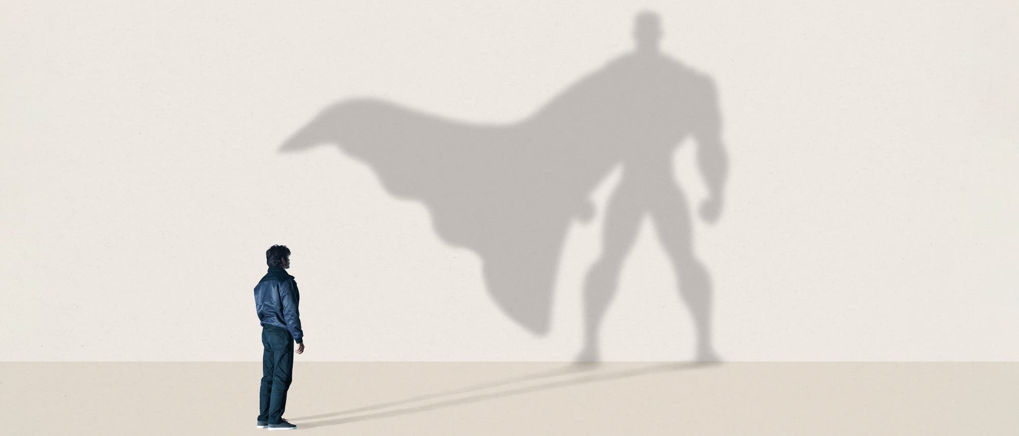 Eine Person steht vor einer Wand, auf der der eigene Schatten als großer Superheld mit Umhang zu sehen ist. Die Szene vermittelt das Wunschdenken von innerer Stärke und Potenzial. Der Hintergrund ist schlicht und hell, wodurch der Schatten deutlich hervorsticht.