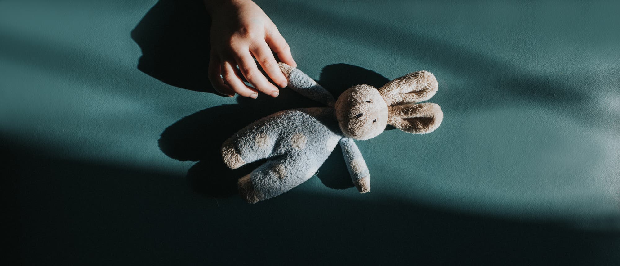 Eine Hand hält ein Plüschtier in Form eines Hasen auf einer dunkelgrünen Oberfläche. Das Bild ist von Schatten durchzogen, die eine ruhige und nachdenkliche Stimmung erzeugen. Der Hase ist aus weichem Stoff und hat lange Ohren.