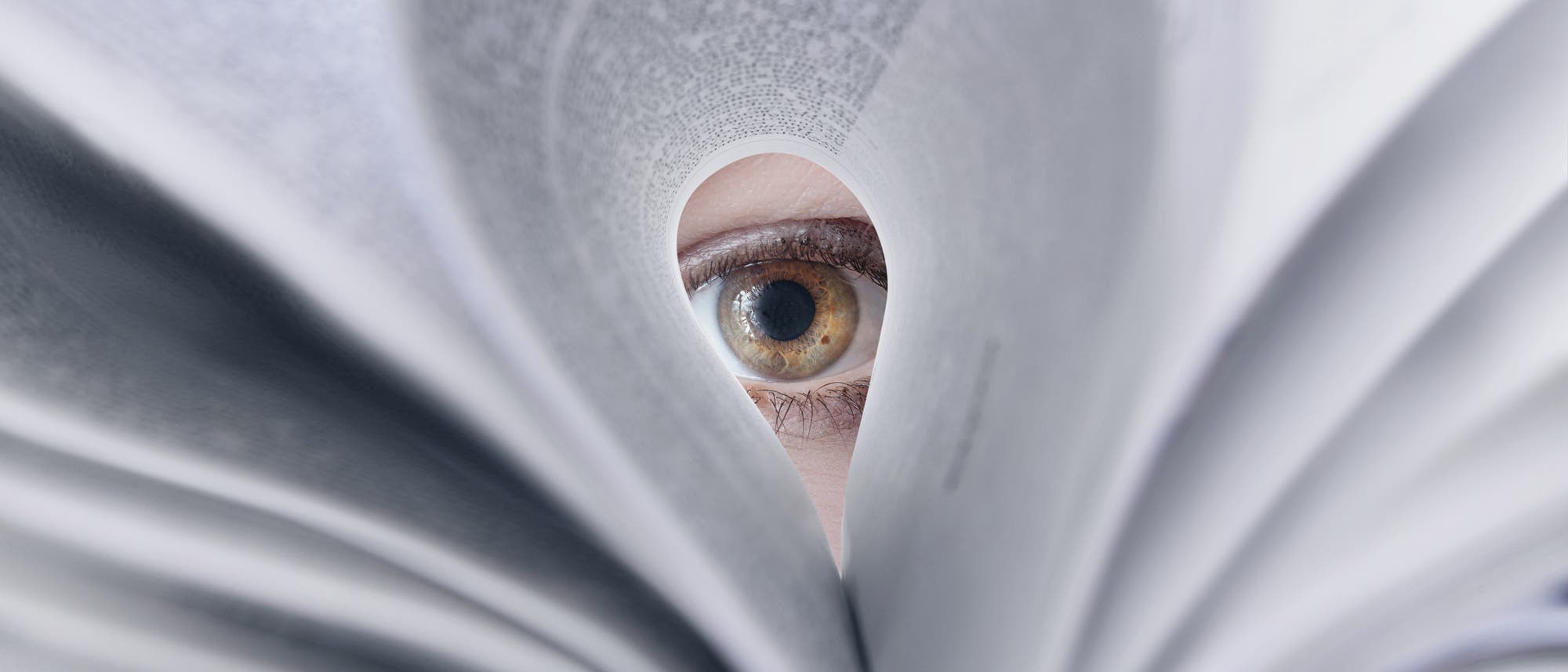 Ein Auge blickt durch eine herzf&ouml;rmige &Ouml;ffnung in einem aufgeschlagenen Buch. Die Seiten des Buches sind unscharf im Vordergrund, w&auml;hrend das Auge im Fokus steht. Das Bild vermittelt den Eindruck von Neugier und Entdeckung.