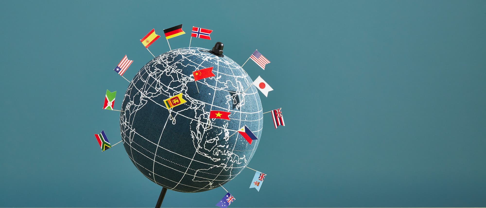 Ein Globus mit eingezeichneten L&auml;ngen- und Breitengraden steht auf einem Tisch. Mehrere kleine Flaggen verschiedener L&auml;nder sind auf der Oberfl&auml;che des Globus verteilt, darunter die Flaggen von Deutschland, China, den USA und Japan. Der Hintergrund ist einfarbig blau. Der Globus symbolisiert internationale Vielfalt und globale Vernetzung.